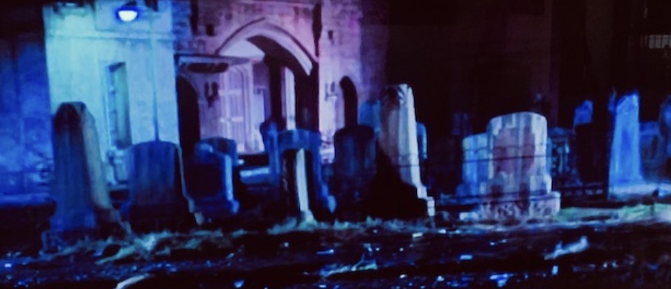 A dimly lit graveyard with old gravestones and a gothic building in the background, illuminated by a faint blue light.