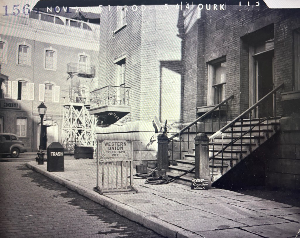 Historic black and white photograph of a street scene featuring a Western Union Telegraph sign, a trash bin, and a staircase leading to a brick building. The background includes lamp posts and an old car.