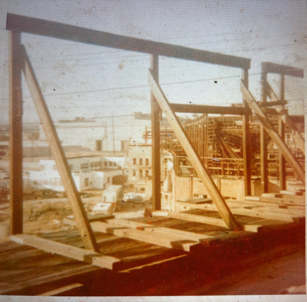 Construction site showing wooden framework and support beams, with a view of buildings and equipment in the background.