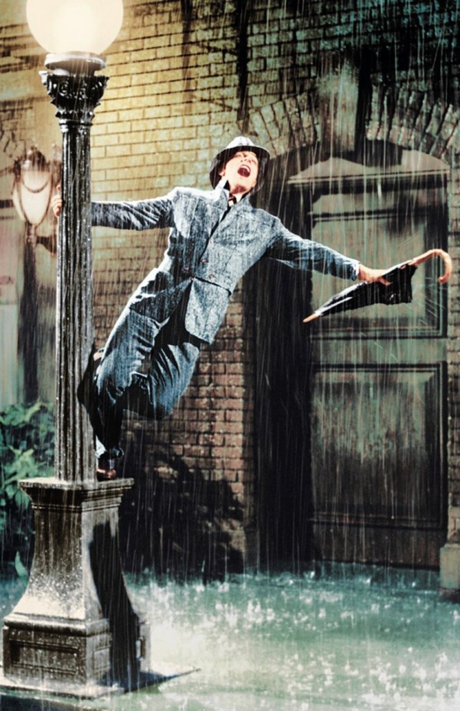 A man in a gray suit and hat joyfully dances and sings while holding an umbrella near a streetlamp, drenched in rain, with water pooling at his feet.