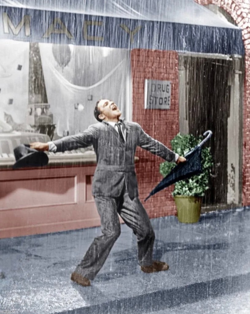 A man in a gray suit joyfully dancing in the rain, holding an umbrella and a hat, in front of a storefront with the sign 'Macy'.
