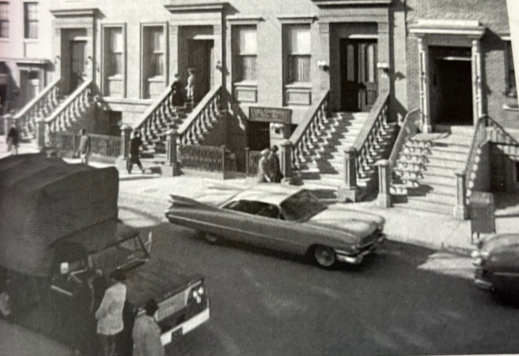 Black and white image of a street scene featuring several brownstone buildings with stairs and railings. People can be seen walking on the sidewalk, and vintage cars are parked on the street.