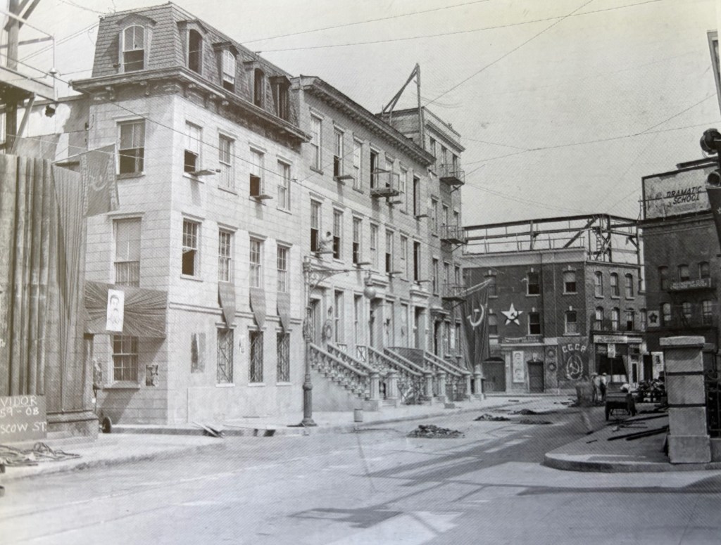 Black and white historical photograph of a street scene featuring buildings with banners and flags, including a Turkish flag and Soviet symbols, along with construction elements visible in the background.