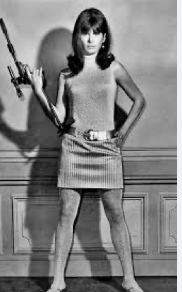 A young woman in a sleeveless top and mini skirt stands confidently with a toy gun in her hand, posing in front of a textured wall.