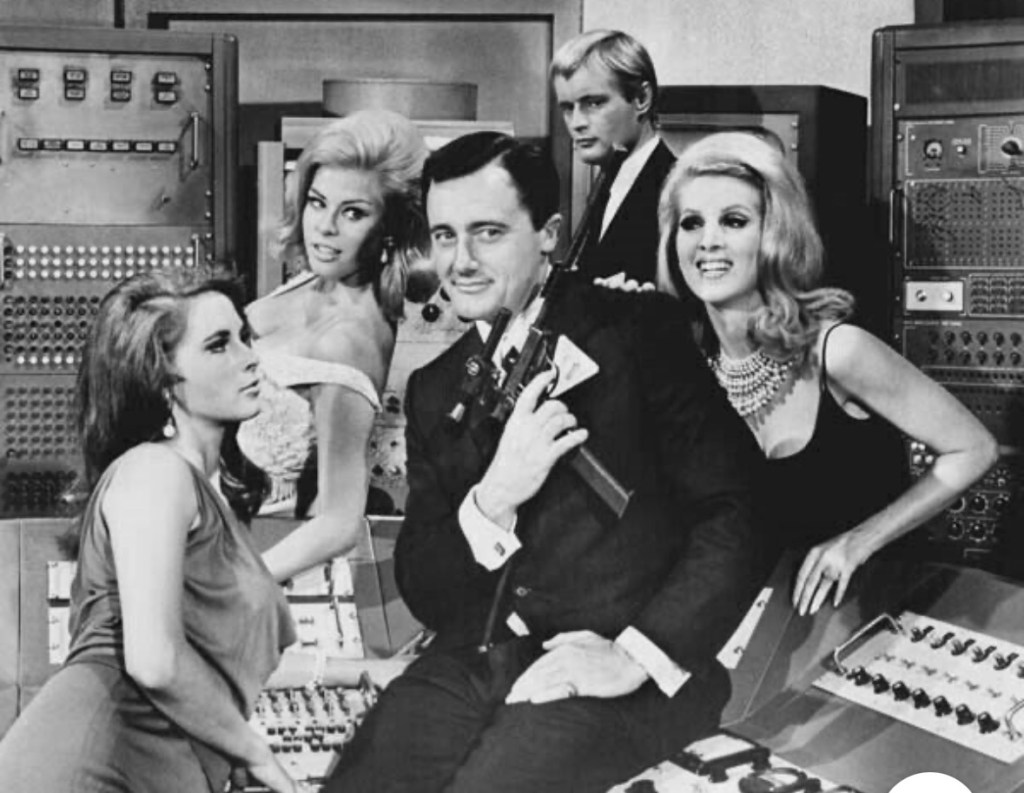 Black and white vintage image featuring a man holding a gun surrounded by four women, posed in a retro control room setting.
