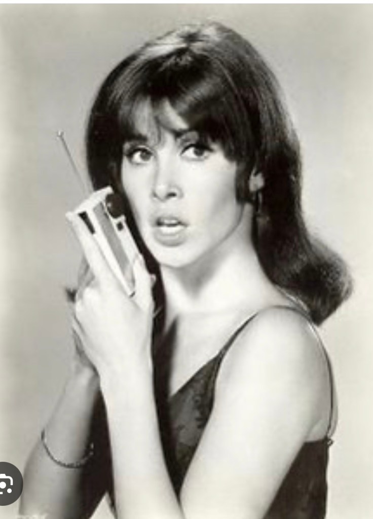 A woman with dark hair and makeup, holding a retro communication device and looking intently at the camera.