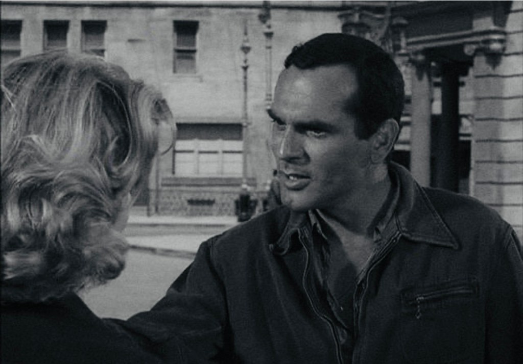 A man and a woman engage in an intense conversation on a city street, with the man looking assertive and the woman appearing concerned.