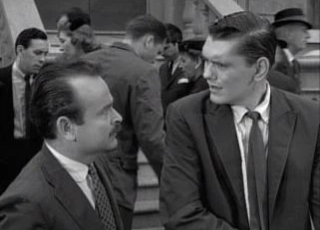 Two men in suits stand facing each other in a crowded outdoor setting, appearing engaged in conversation, with several people walking in the background.