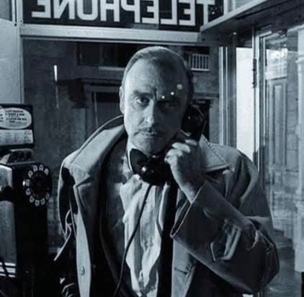 A man in a trench coat holding a rotary phone inside a phone booth, with a sign reading 'TELEPHONE' visible in the background.