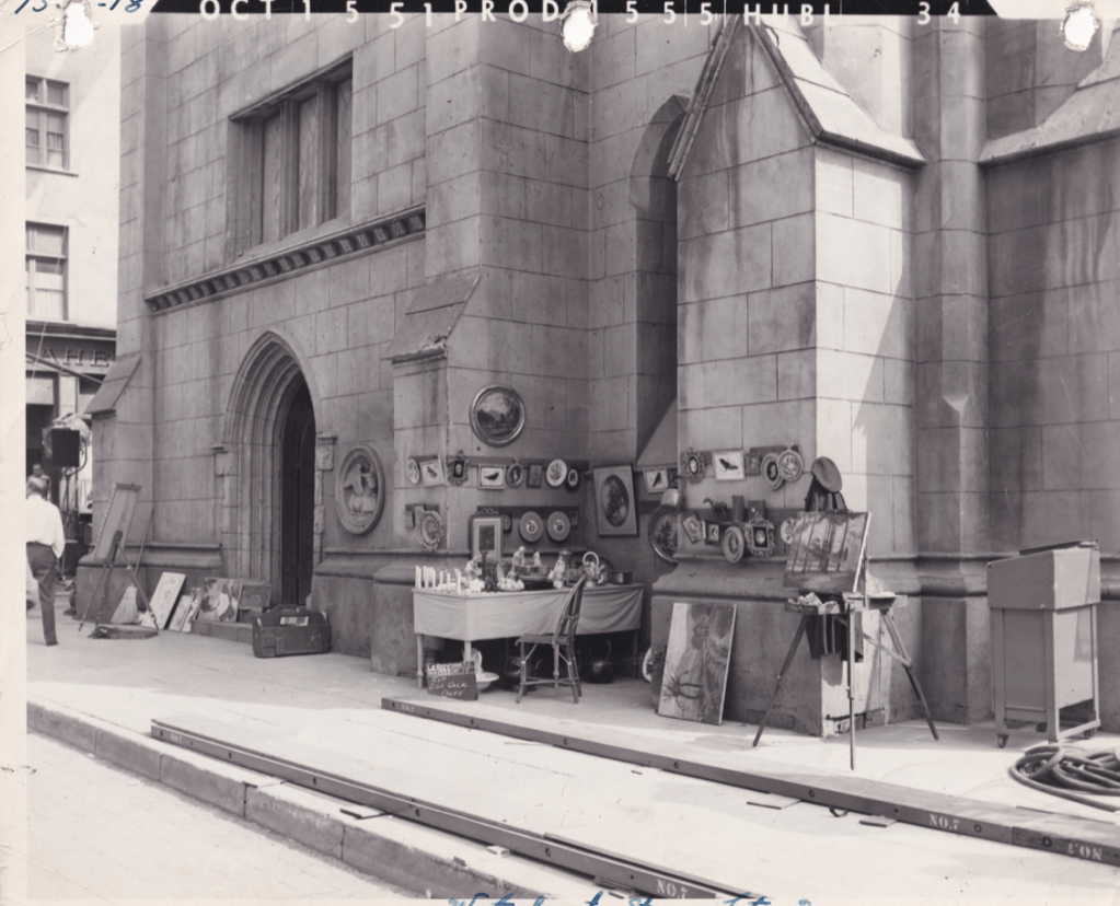 A historical photograph showing a stone building facade with an arched entrance. Near the entrance, there are tables displaying various items, including paintings and decorative plates. The scene includes a person walking by and a couple of artists' easels.