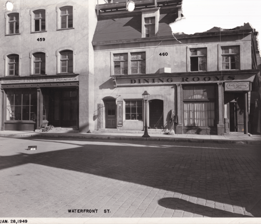 Historic black and white photograph of two buildings on Waterfront Street, labeled '459' and '460'. The left side features an upholstery store named 'Tribby's Polishers' with large windows, while the right has a dining establishment with a sign reading 'Dining Rooms' and a 'Early Morning Café' sign.