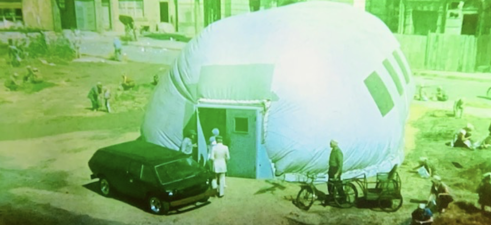 A large, inflatable dome structure with a blue door, surrounded by people engaged in outdoor activities. A parked black vehicle is seen in front of the dome, and bicycles are nearby.